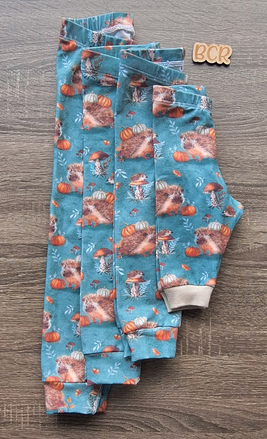 Four pairs of handmade leggings folded in half. The fabric features hedgehogs, pumpkins, mushrooms, leaves on a jadeite green background. The leggings are made using organic cotton.